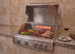 Churrasqueira a gás Evol em aço inox com queimador infrared traseiro em funcionamento, instalada em bancada de pedra. Grelha com carnes, legumes e frango sendo preparados, destacando o calor radiante para finalização e douramento dos alimentos.