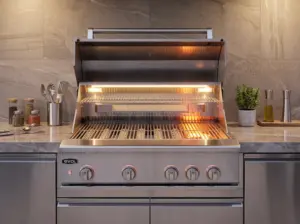 Churrasqueira a gás Evol em aço inox com tampa aberta, equipada com sear burner, grelhas em aço inox e iluminação interna em LED. Instalada em bancada planejada de área gourmet, com utensílios e temperos ao fundo, destacando a ampla área de preparo para carnes e grelhados.