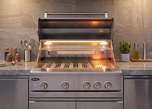 Churrasqueira a gás Evol em aço inox com tampa aberta, equipada com sear burner, grelhas em aço inox e iluminação interna em LED. Instalada em bancada planejada de área gourmet, com utensílios e temperos ao fundo, destacando a ampla área de preparo para carnes e grelhados.