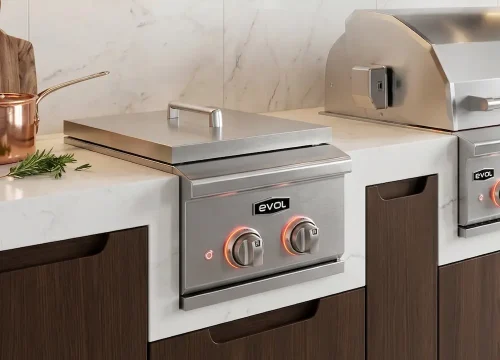 Side burner embutido em aço inox integrado à bancada de área gourmet planejada, com dois queimadores de alta potência, instalado ao lado de uma churrasqueira a gás Evol para preparo simultâneo de acompanhamentos e finalizações.